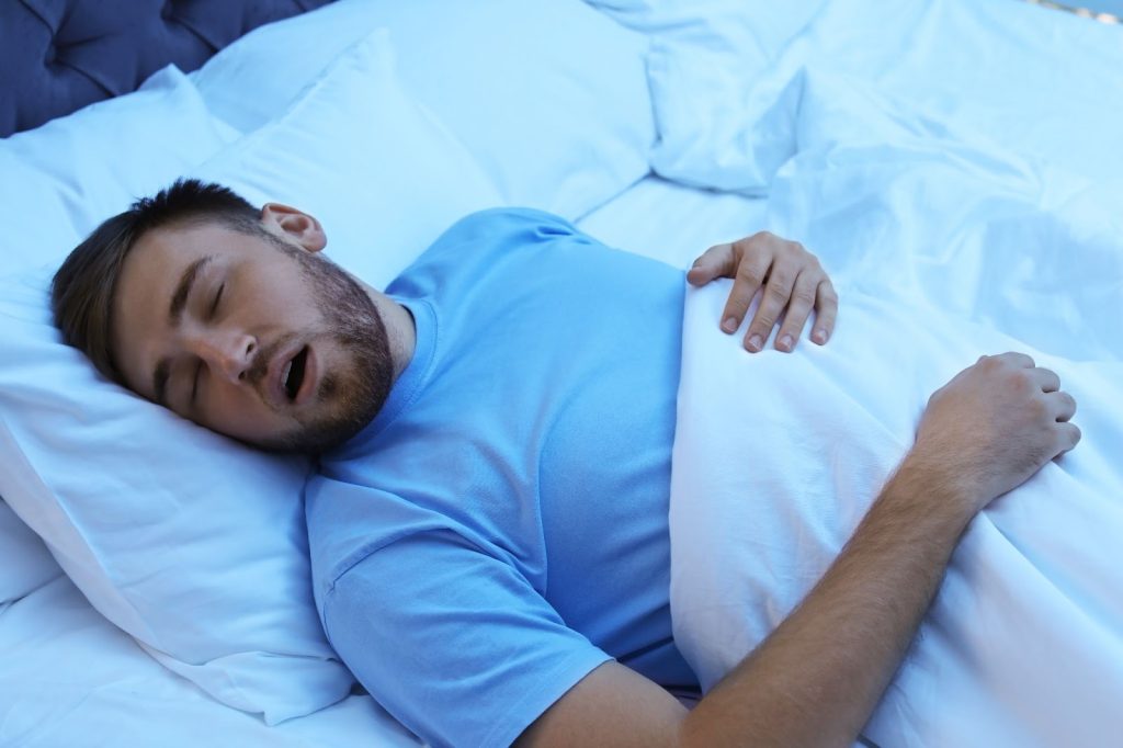 Man sleeping on bed in blue shirt with mouth open under white blanket.