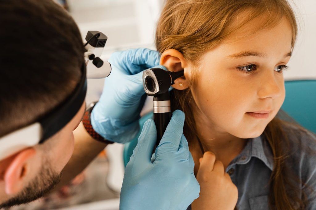 A close-up of a doctor wearing blue gloves and a headlamp using an otoscope to perform an ear examination on a young girl.