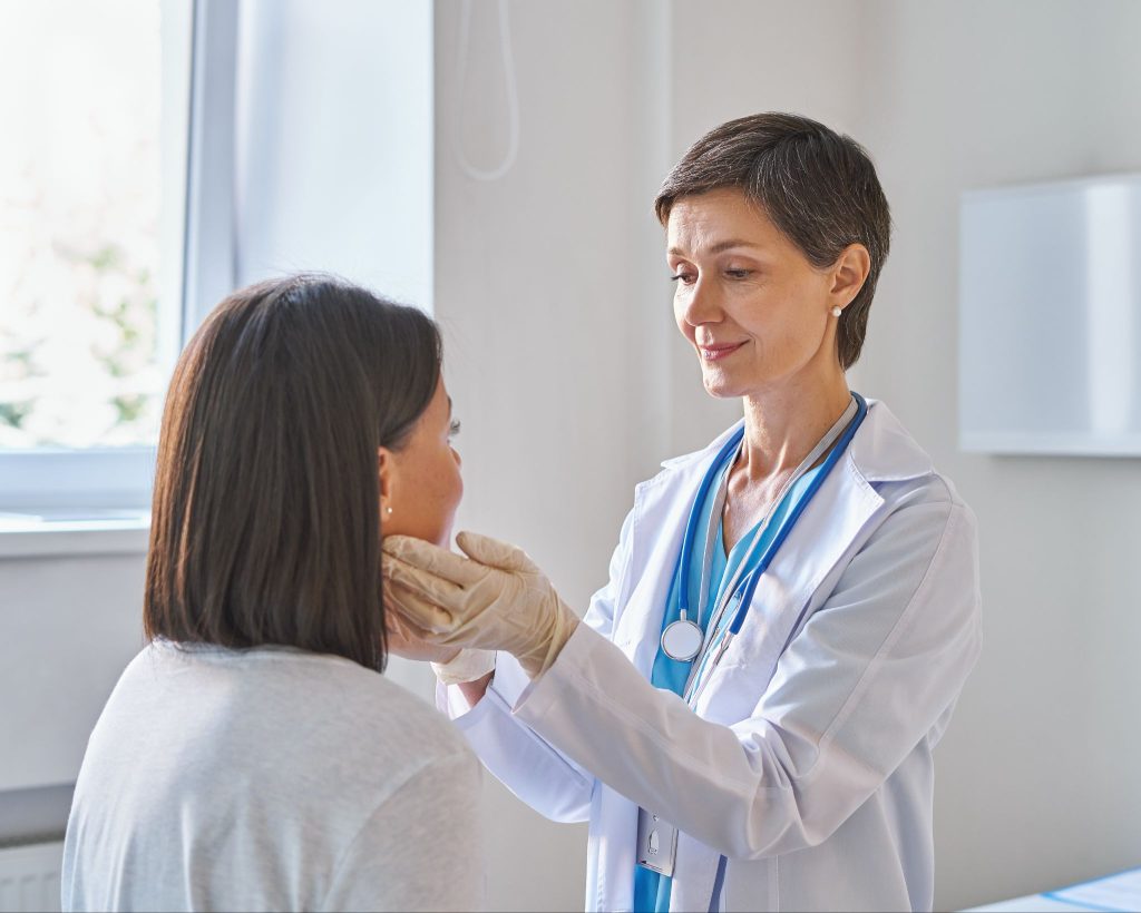 A female doctor performing a physical examination of a patient's neck and lymph nodes in a clinical setting.