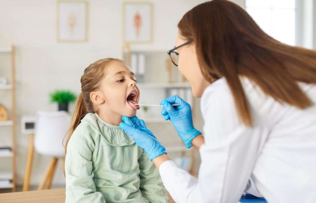 A female doctor wearing blue gloves uses a tongue depressor to examine the throat of a young girl in a clinic setting.