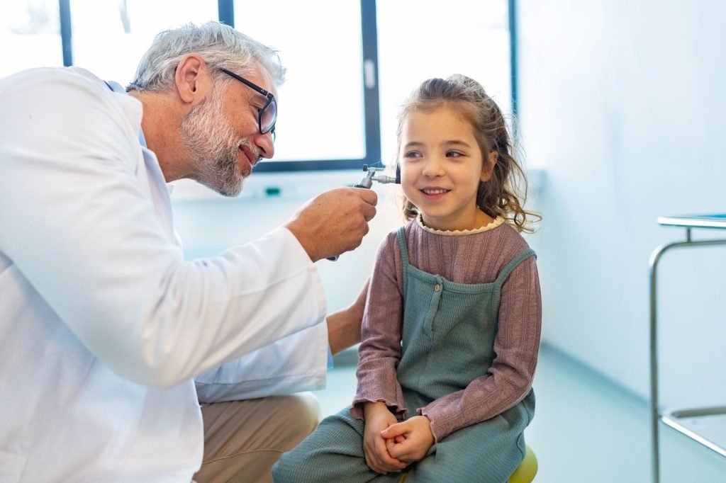 A senior male doctor using an otoscope to examine the ear of a smiling young girl during a medical checkup.