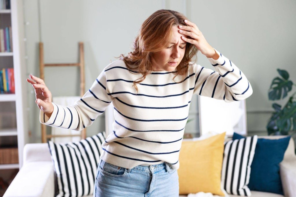 A woman stands in a living room holding her head in pain expressing discomfort and distress