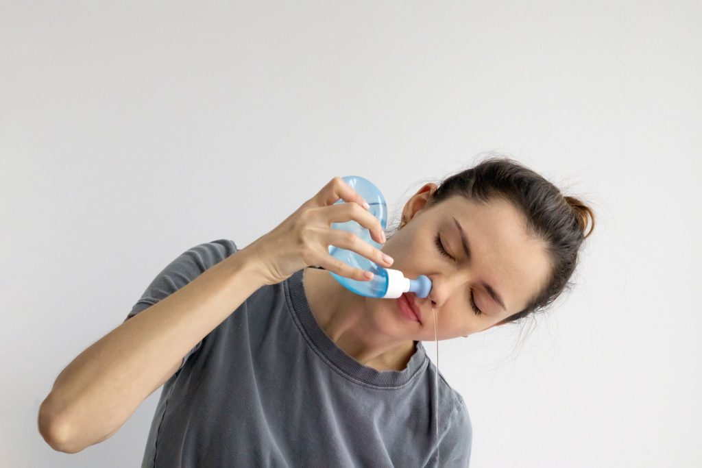 Woman using a blue neti pot to perform a nasal saline rinse against a plain white background.