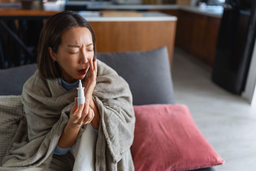 Woman wrapped in a blanket sitting on a couch holding a nasal spray bottle while touching her cheek in discomfort.
