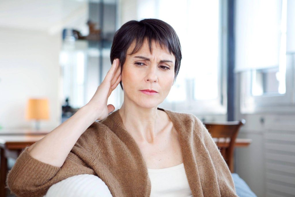A middle aged woman in a brown cardigan presses her hand to her temple looking worried and uncomfortable