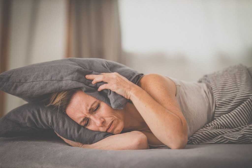 A weary woman lies curled in bed clutching a gray pillow over her head and grimacing