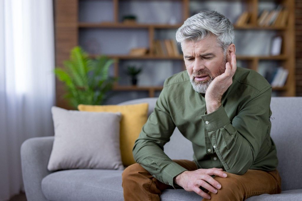 A worried middle aged man sits on a living room sofa pressing a hand to his ear as if in pain