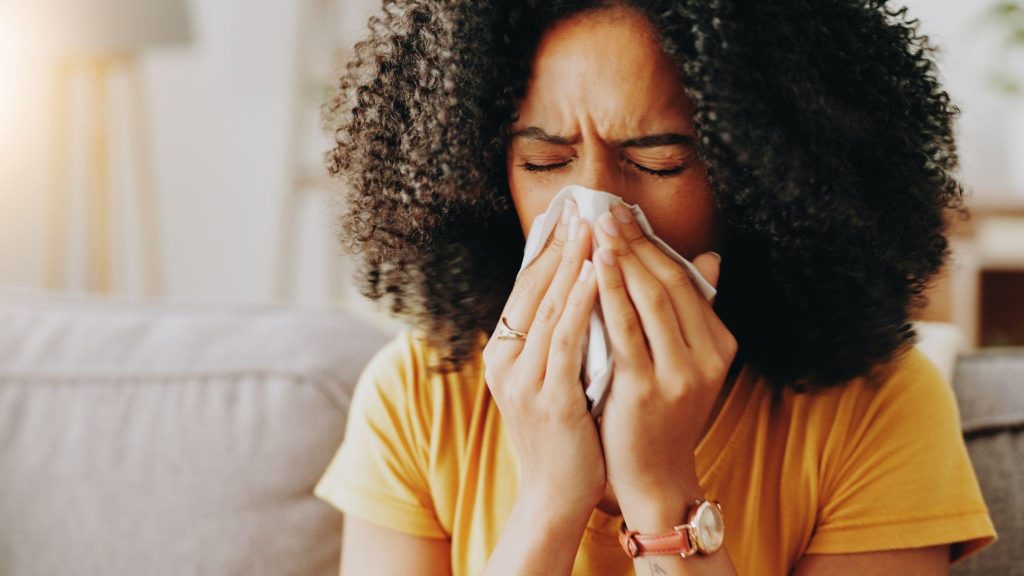 A woman sneezes on a couch reflecting her allergy symptoms with tissues placed beside her for relief